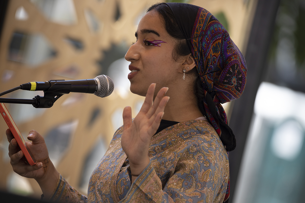 Woman dressed in patterned headscarf and dress stands at a microphone, gesticulating with one hand and holding a smartphone with the other. She wears sparkling purple eyeliner.